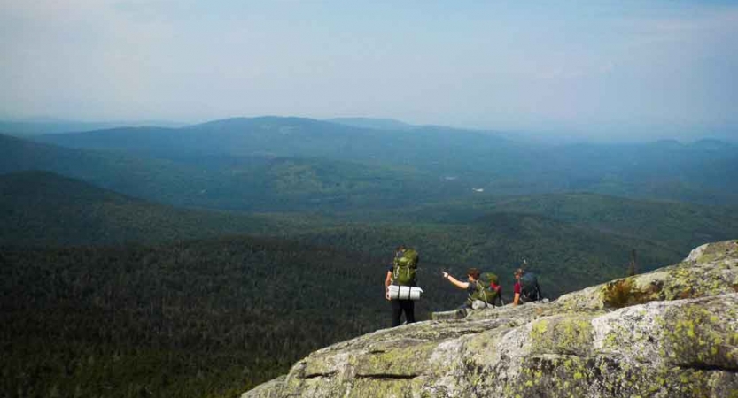 three people wearing backpacks rest on a rocky overlook with a green mountainous landscape in the background